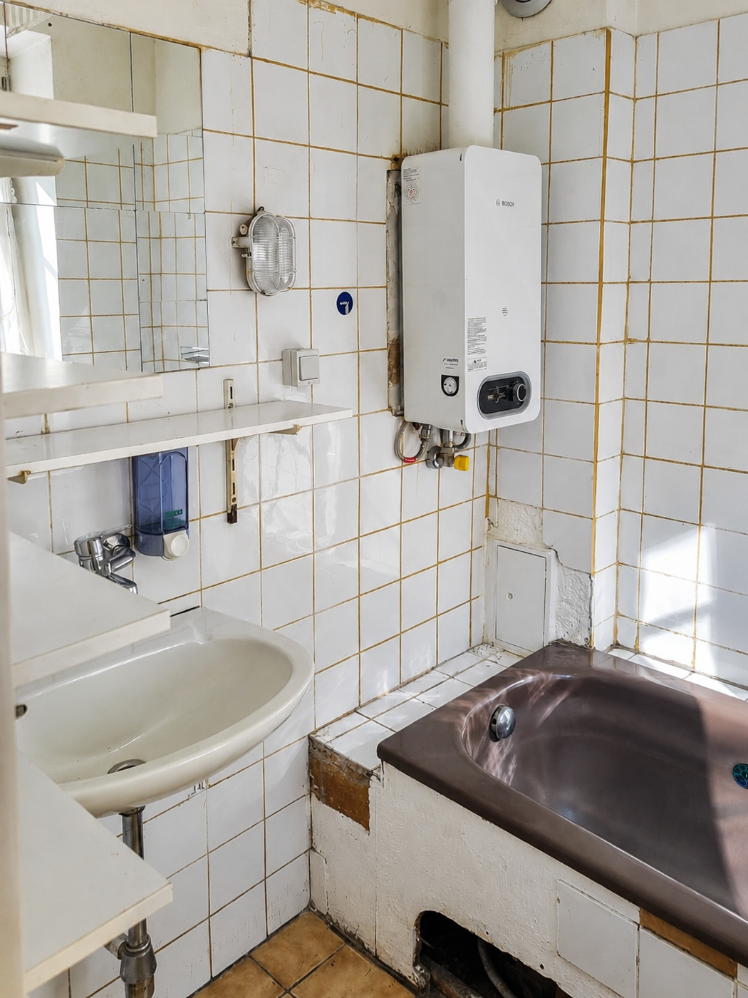 Älteres kleines Bad mit Wanne, verfärbten Fliesen und offen montierter Therme